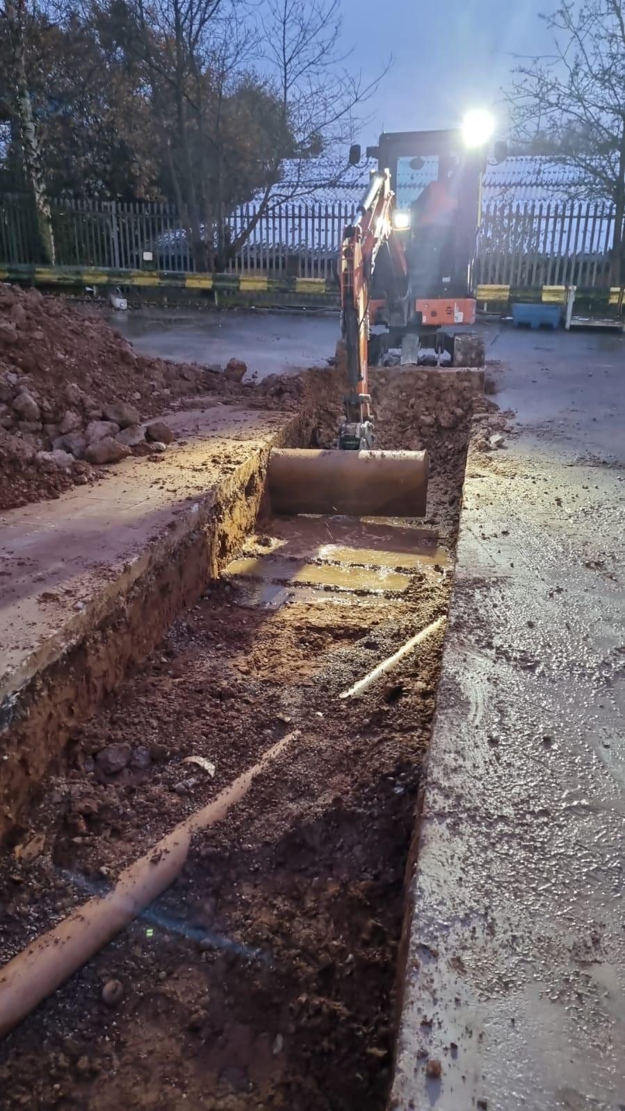 Excavator digging a drainage trench beside a concrete slab at dusk